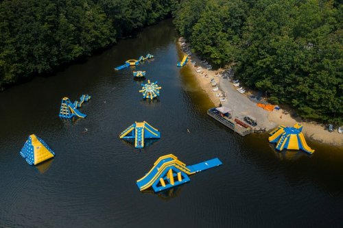 Aerial view of a water park with inflatable structures on a river, surrounded by trees and a sandy beach area.