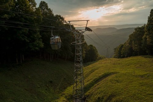 A cable car glides over a green hillside at sunset, surrounded by trees and distant mountains.