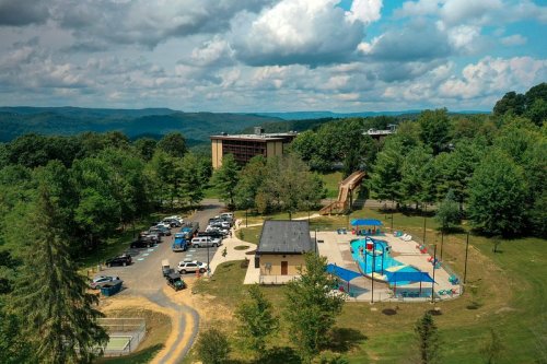 Aerial view of a pool area with slides, surrounded by trees and mountains, with parked cars nearby.