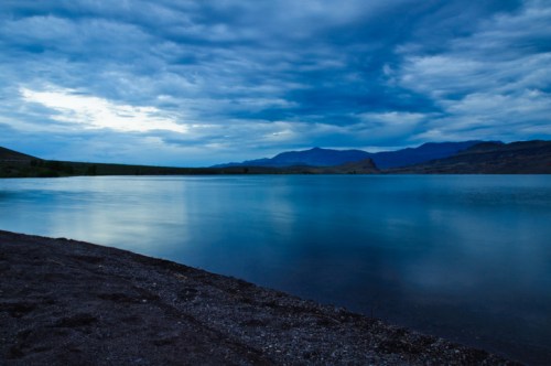 A tranquil lake at dusk, reflecting blue skies and clouds, with a dark shoreline in the foreground.