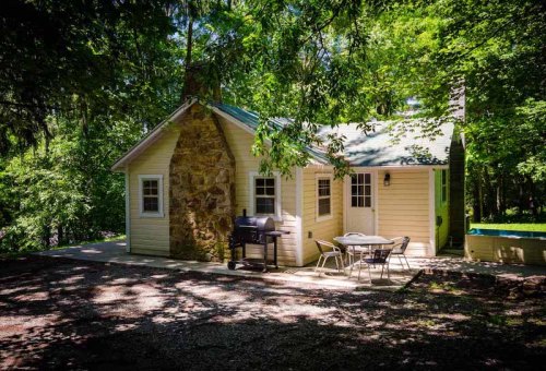 A cozy cabin surrounded by trees, featuring a stone chimney, outdoor seating, and a grill on a gravel path.