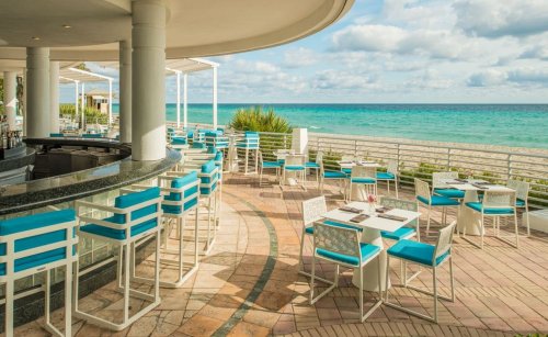 Outdoor dining area with turquoise chairs overlooking a beach and ocean under a partly cloudy sky.