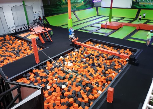 A vibrant indoor trampoline park featuring a foam pit filled with orange and black blocks, surrounded by trampolines.