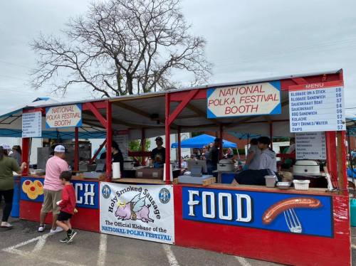 A colorful food booth at a festival, featuring a menu with various items like kielbasa, sandwiches, and drinks.