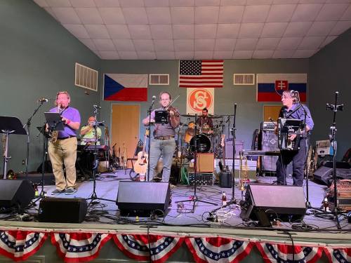 A band performs on stage with instruments and microphones, decorated with flags and patriotic bunting in the background.