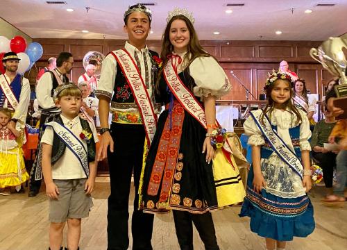 A group of children in traditional costumes, with a boy and girl wearing crowns and sashes, at a festive event.