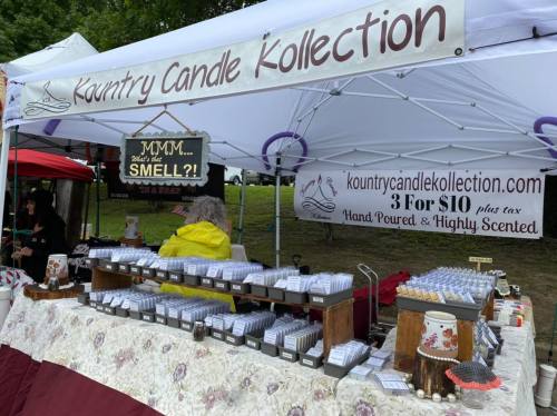 A vendor booth at an outdoor market featuring candles, with a sign reading "MMM... What's that SMELL?!" and promotional offers.