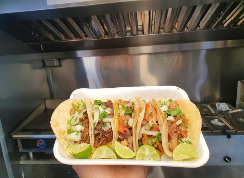 Three tacos on a white plate, filled with meat, onions, and tomatoes, garnished with lime wedges, in a kitchen setting.