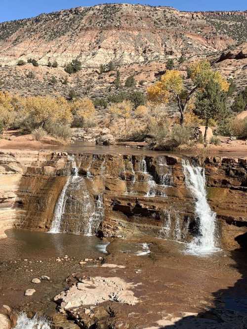 A scenic waterfall cascades over rocky terrain, surrounded by autumn trees and a mountainous landscape.