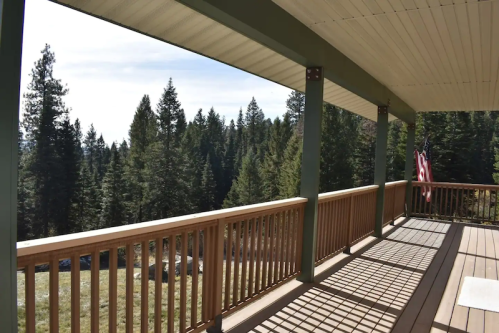 A wooden porch with a railing overlooks a forest of tall pine trees under a clear sky. An American flag is visible.