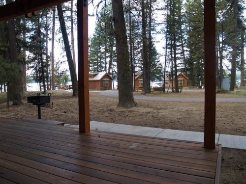 View from a wooden porch overlooking a path, cabins, and trees by a lake.