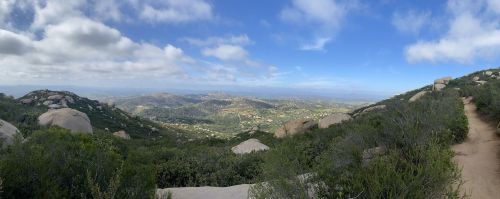 A panoramic view of rolling hills and valleys under a partly cloudy sky, with rocky terrain and greenery in the foreground.