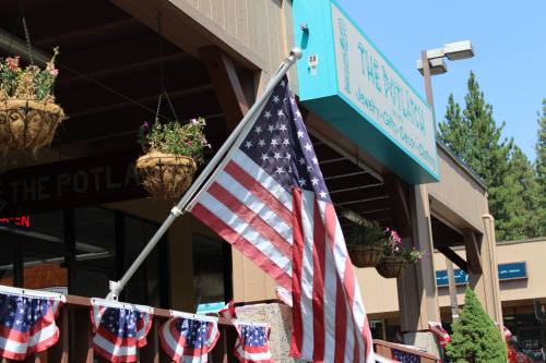 A U.S. flag hangs outside a building with flower baskets and festive decorations, under a clear blue sky.