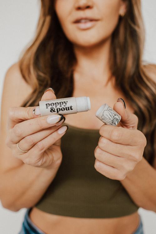 A woman holds a Poppy & Pout lip balm in one hand and its cap in the other, smiling at the camera.