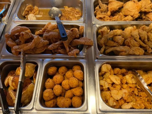 A variety of fried foods in metal trays, including chicken pieces, crispy pork rinds, and golden-brown balls.