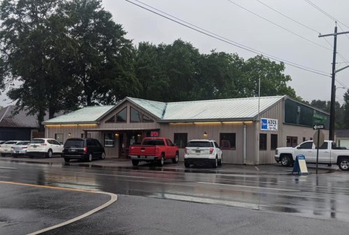 A one-story building with a metal roof, surrounded by parked cars, on a rainy street. "Open" sign visible in the window.