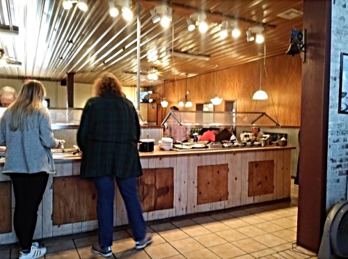 Two people serve themselves at a buffet in a rustic restaurant with wooden decor and warm lighting.