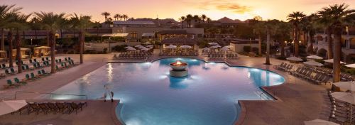A serene pool area at sunset, surrounded by palm trees and lounge chairs, with a fire pit in the center.