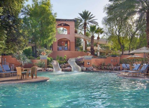 A serene pool area with water slides, surrounded by palm trees and lounge chairs in a sunny resort setting.