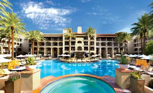 A luxurious hotel with a large pool surrounded by palm trees and lounge chairs under a clear blue sky.