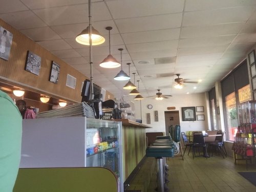 A cozy diner interior with green and blue chairs, wooden accents, and pendant lights, featuring a counter and tables.