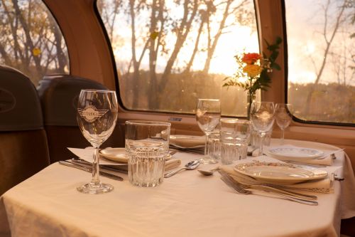 A beautifully set dining table in a train car, with glassware, plates, and a flower vase, against a sunset backdrop.