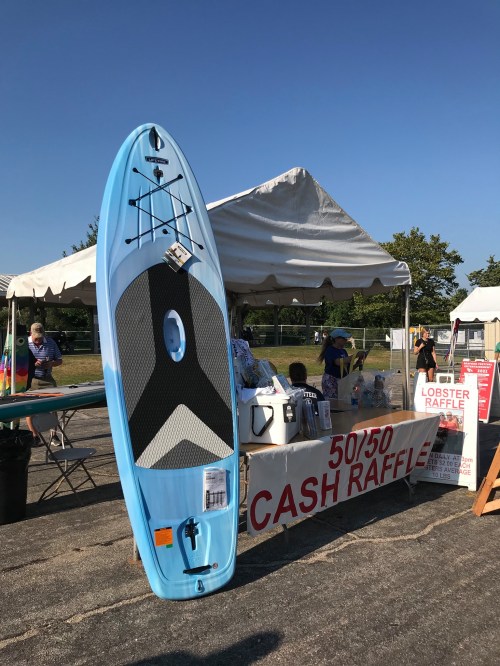 A blue paddleboard stands next to a booth with raffle signs and various items at an outdoor event.