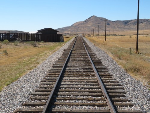 A view of railway tracks stretching into the distance, flanked by dry grass and a distant mountain under a clear blue sky.