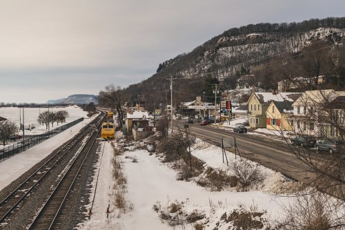 A snowy landscape with train tracks, a road, and small buildings alongside a hillside under a cloudy sky.