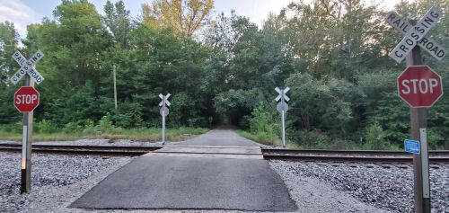 A quiet railroad crossing with stop signs, surrounded by trees and gravel, leading to a path in the distance.