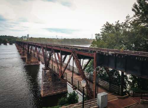 A rusted metal bridge spans a calm river, surrounded by trees and a cloudy sky.