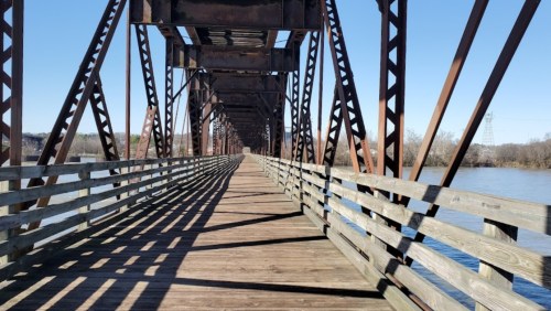 A wooden bridge with metal supports stretches over a calm river, under a clear blue sky. Shadows create patterns on the walkway.