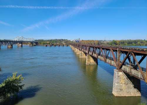 A rusted railway bridge spans a wide river under a clear blue sky, with greenery along the banks and another bridge in the distance.