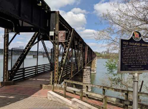 A historic railroad bridge spans a river, with a sign and trees in the background under a partly cloudy sky.