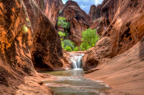 A serene waterfall cascades into a pool surrounded by towering red rock formations and lush greenery.
