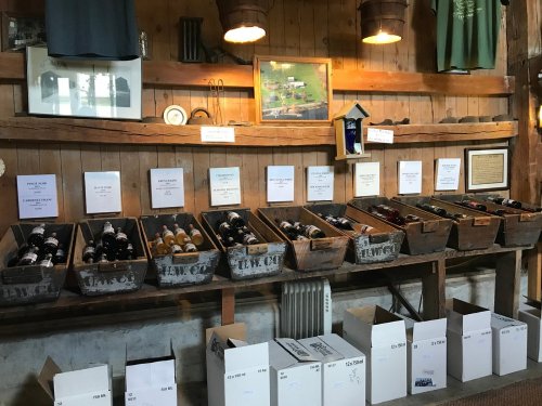 A rustic wooden wall displays wine bottles in crates, with labels and boxes for packaging below.