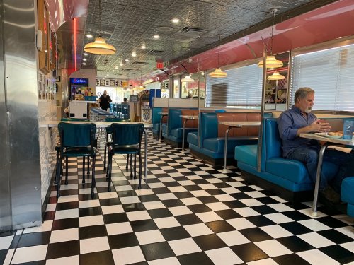 A retro diner interior with checkered floors, blue booths, and a man sitting at a table. Bright lighting and decor.