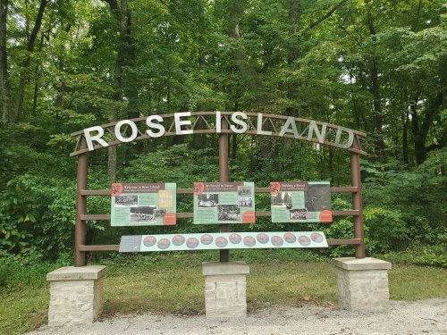 Sign for Rose Island with historical information panels, surrounded by lush green trees.
