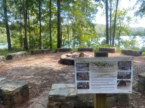 A stone fire pit area surrounded by trees, with a sign detailing the site, overlooking a lake in the background.