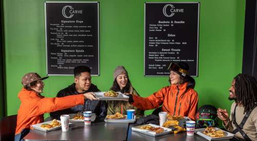 A group of four friends enjoying food at a restaurant with a green wall and menu boards in the background.