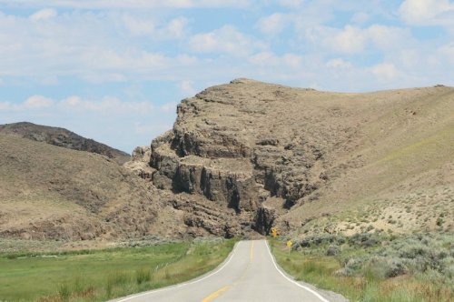 A winding road leads through a rocky landscape with hills under a blue sky and scattered clouds.