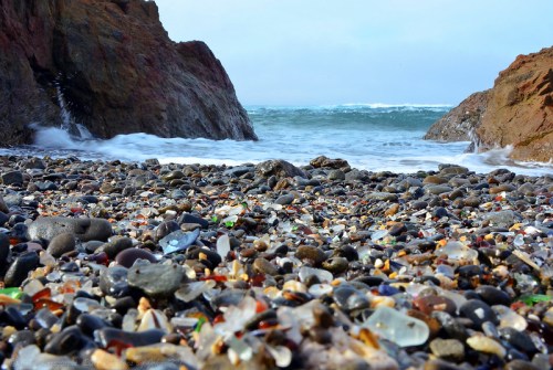 A rocky beach covered in colorful sea glass and pebbles, with waves crashing in the background.
