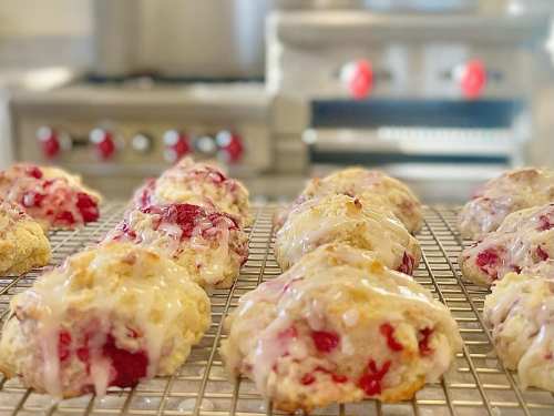 Freshly baked scones with a glaze, featuring bits of red fruit, cooling on a wire rack in a kitchen.