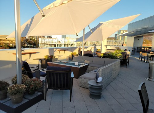 A rooftop lounge area with seating, umbrellas, and plants, overlooking a cityscape in the background.