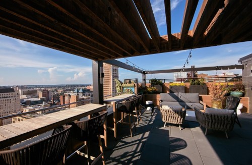 Rooftop terrace with seating, wooden beams, and city skyline views under a clear blue sky.