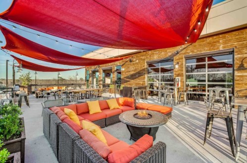 Outdoor seating area with red canopies, cozy couches, and a fire pit, surrounded by wooden walls and tables.