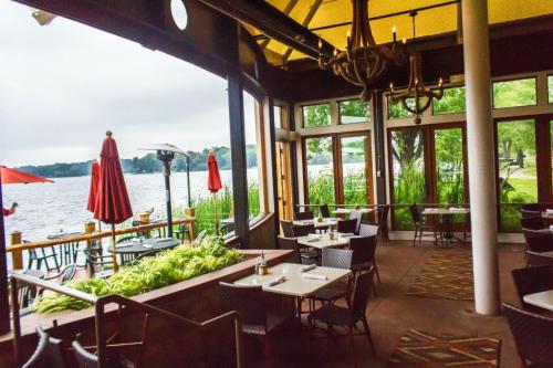 Cozy restaurant interior with tables, overlooking a lake through large windows, surrounded by greenery and umbrellas.