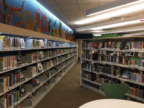 A library interior featuring shelves of books, colorful wall art, and a round table in a well-lit space.