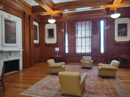 A cozy room with wooden walls, three beige chairs, a rug, and a fireplace, featuring large windows and artwork.