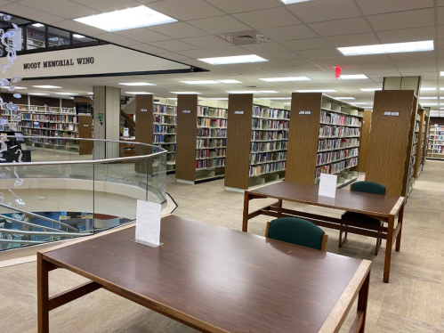 Interior of a library featuring bookshelves, study tables, and a staircase, with a sign reading "Moody Memorial Wing."